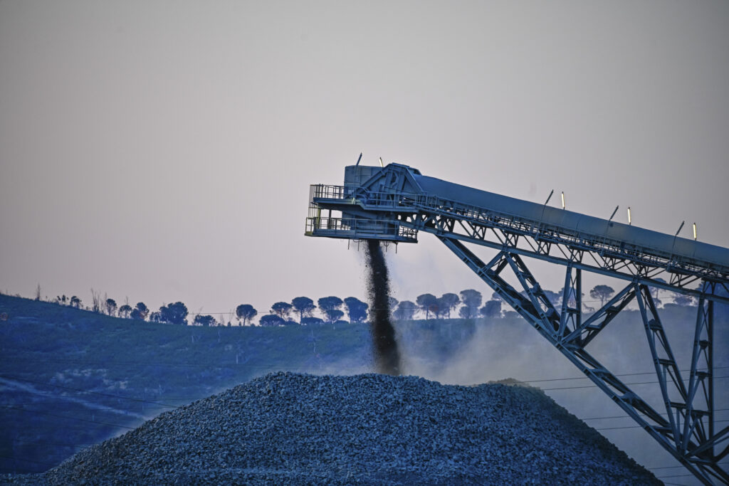 A conveyor belt system pours gravel into a large pile during twilight, with a silhouetted treeline in the background.